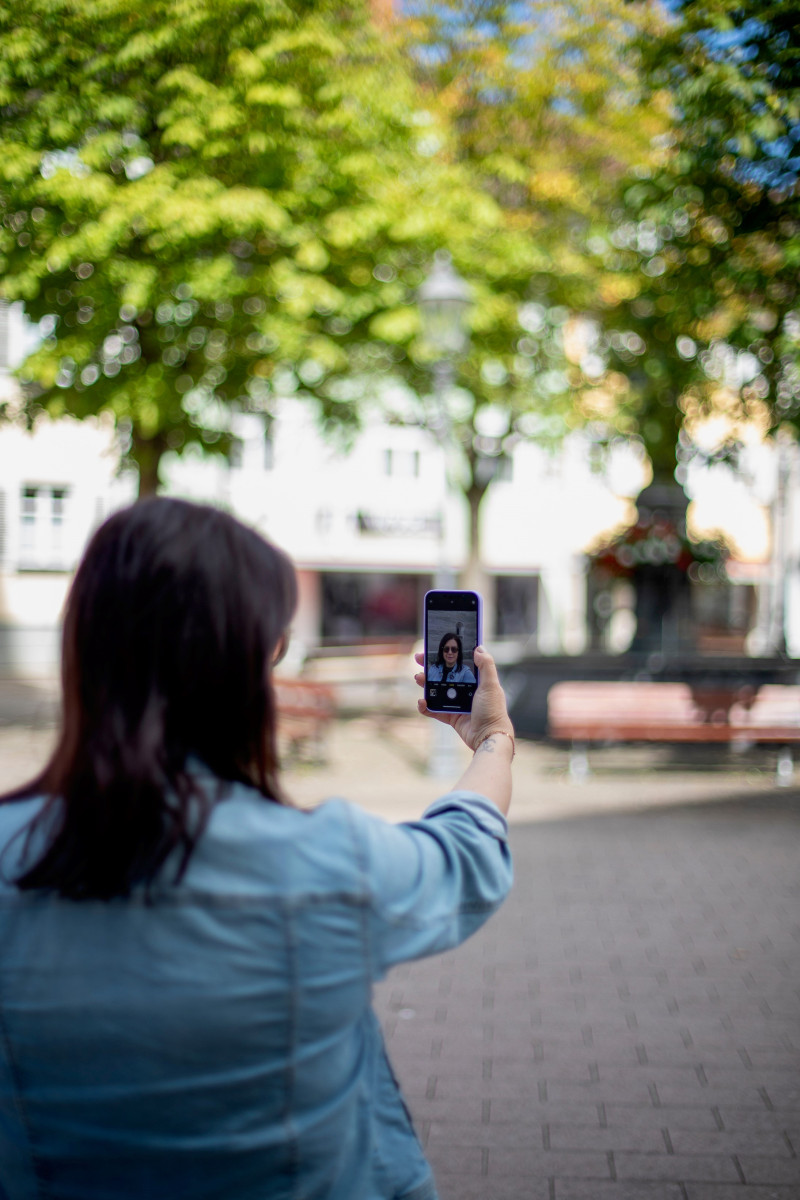 Frau macht Selfie auf Stadtplatz. Smartphone-Fotografie im Urlaub. Brunnen und Bäume im Hintergrund.