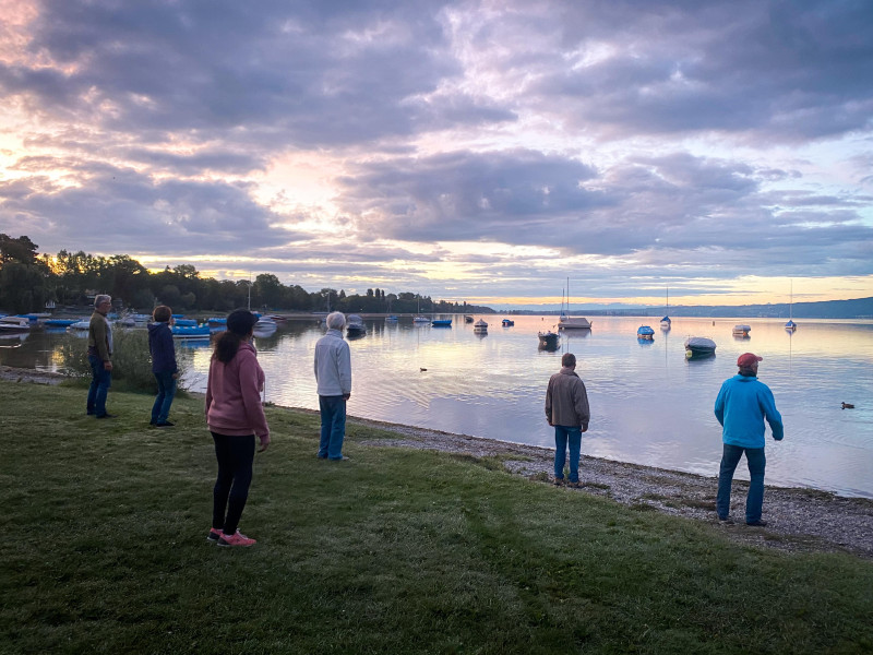 Gruppe am Seeufer bei Sonnenaufgang. Boote auf dem Wasser unter bewölktem Himmel.