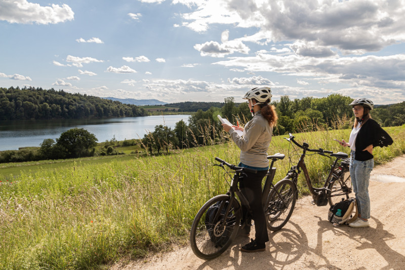 Zwei Frauen mit Fahrrädern auf einem Feldweg mit Blick auf einen See. Radtour mit Karte und E-Bikes.