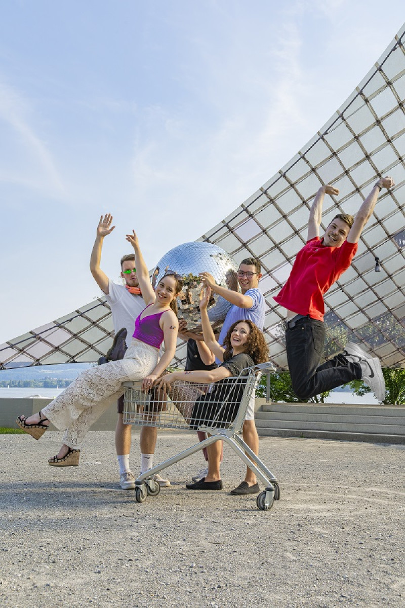 A group of five joyful friends with a disco ball, one jumping, two in a shopping cart, posing in front of a glass building.