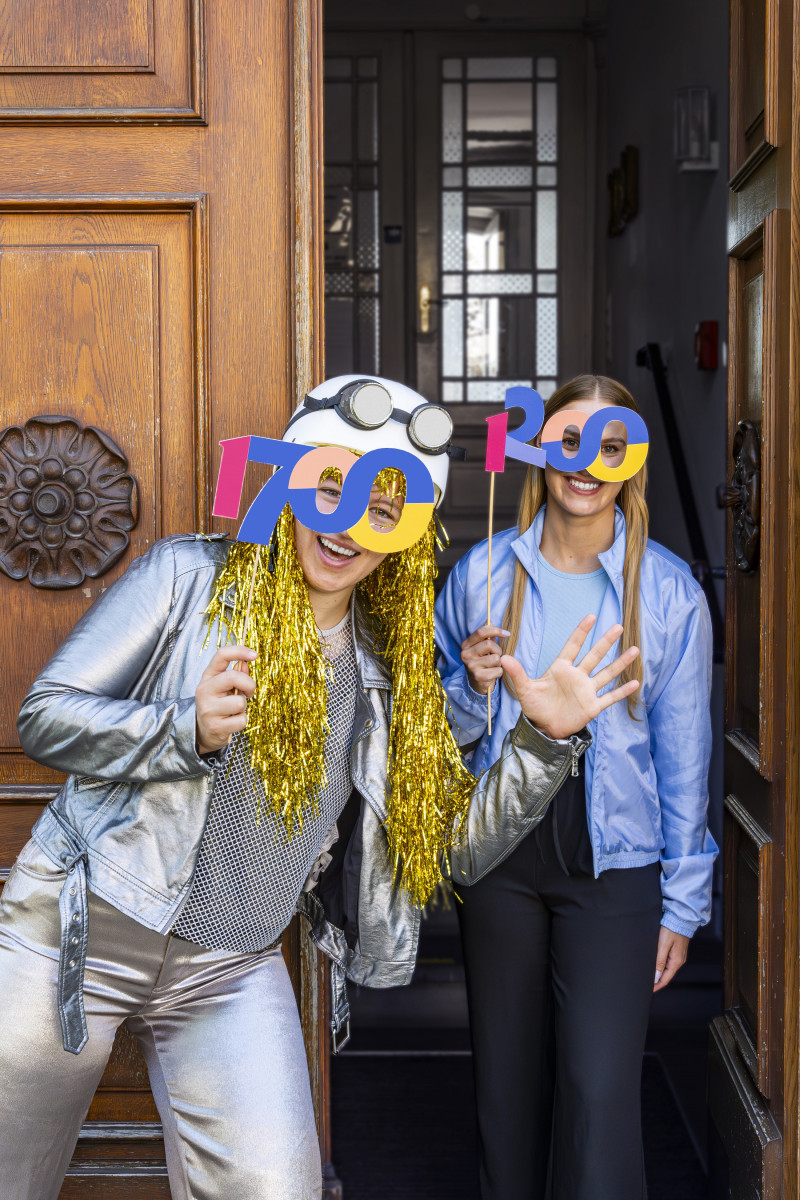 Zwei junge Frauen feiern mit bunten 700-Jahre-Jubiläums-Brillen vor einer Tür. Festliche Stimmung und stilvolle Kleidung.