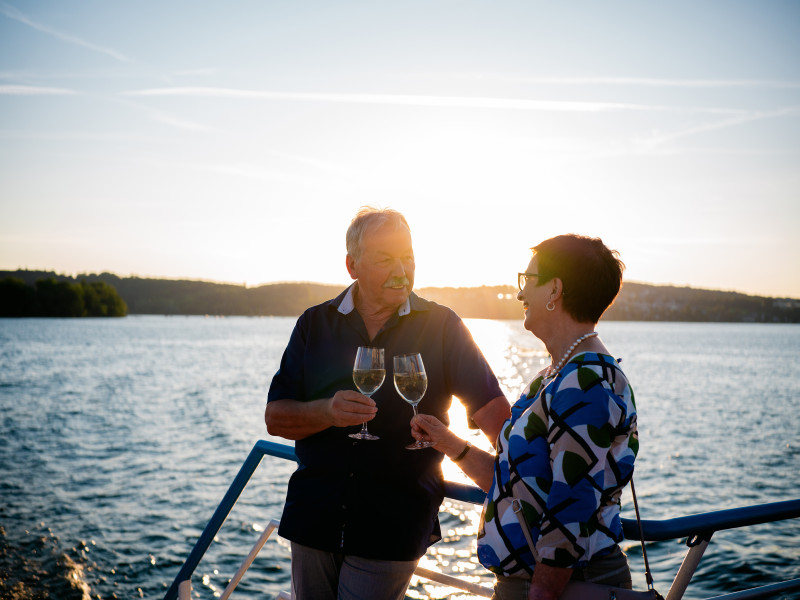 Älteres Paar stößt auf einem Boot mit Wein an, Sonnenuntergang im Hintergrund. Romantische Kreuzfahrt.